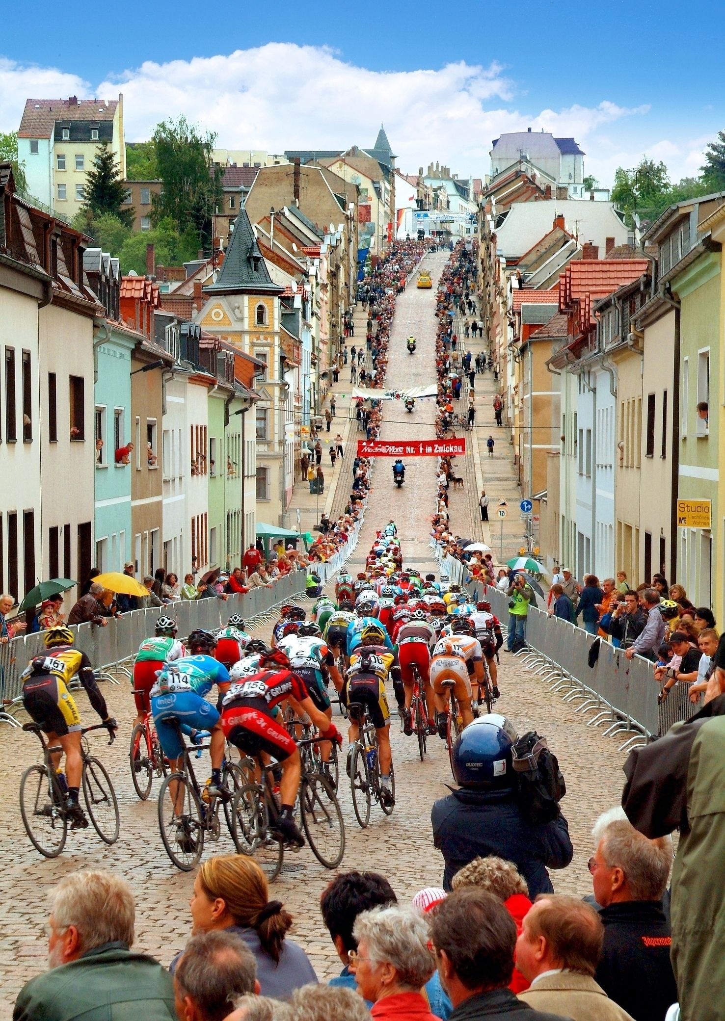 Una calle adoquinada flanqueada por edificios coloridos, una multitud de ciclistas y espectadores siguiendo una carrera de ciclismo.