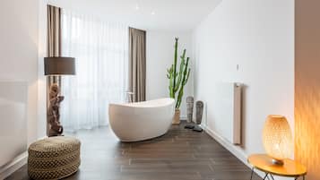 A modern bathroom with a freestanding bathtub, a round ottoman, a floor lamp, and a tall cactus plant.