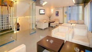 Un moderno vestíbulo de hotel con sofás de cuero blanco, un separador de vidrio, una recepción y un televisor montado.