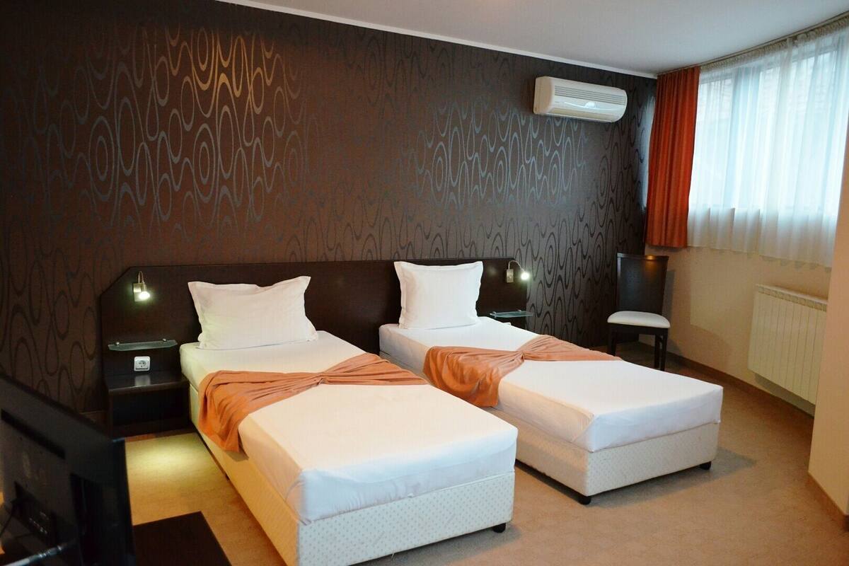 A hotel room with two beds, a wall-mounted air conditioner, a bedside table with a lamp, and a window with curtains.