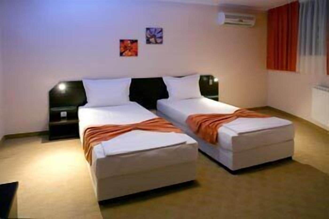 Une chambre double avec deux lits, chacun étant équipé d’un oreiller et d’une couverture. Il y a deux tables de chevet avec des lampes. La chambre a un sol recouvert de moquette et une fenêtre avec des rideaux.