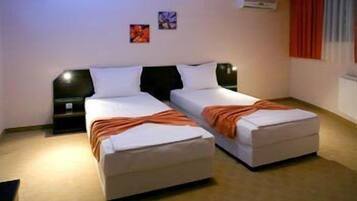Une chambre double avec deux lits, chacun étant équipé d’un oreiller et d’une couverture. Il y a deux tables de chevet avec des lampes. La chambre a un sol recouvert de moquette et une fenêtre avec des rideaux.