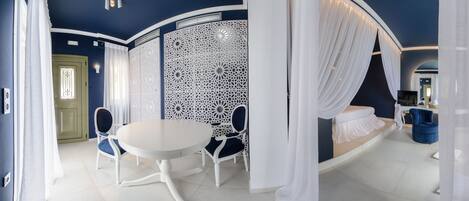 A room with a round table, two chairs, a white partition with intricate designs, a bed with white curtains, and a blue accent wall.