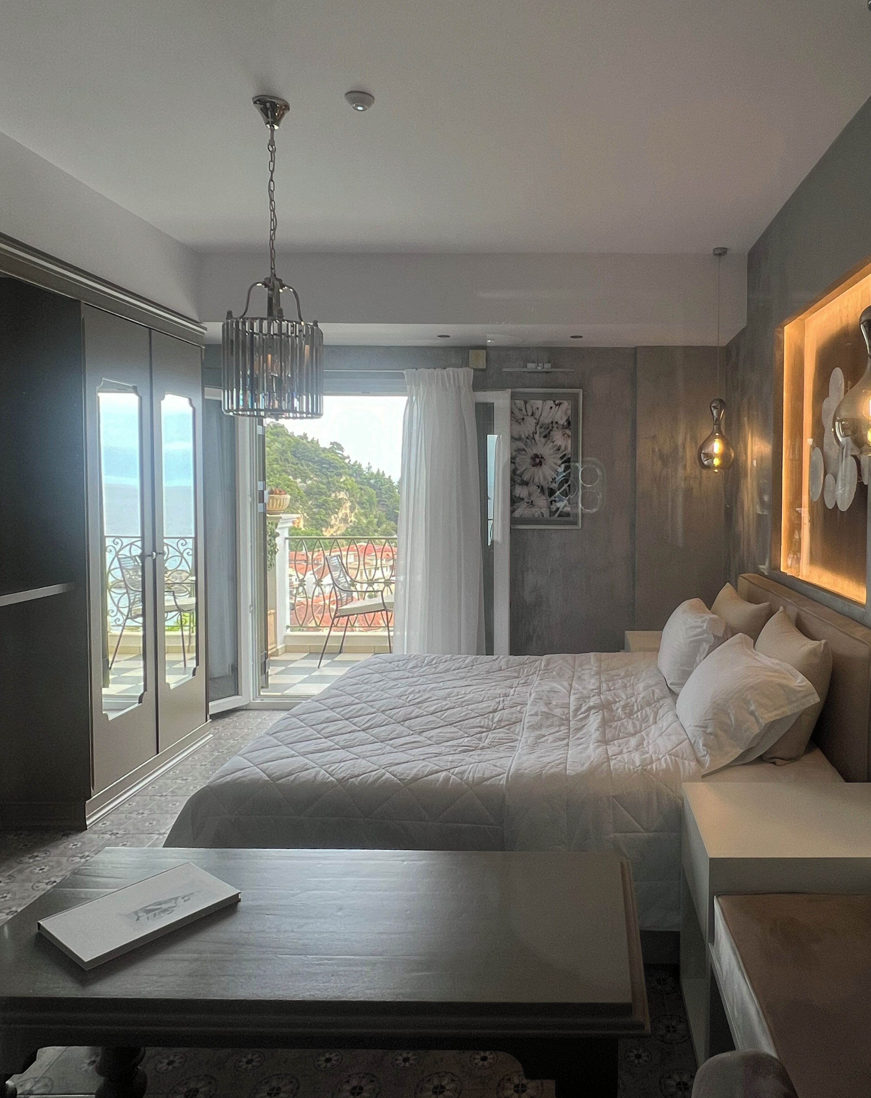 A hotel room with a bed, a bedside table, a sofa, a balcony with a view, and a ceiling light fixture.