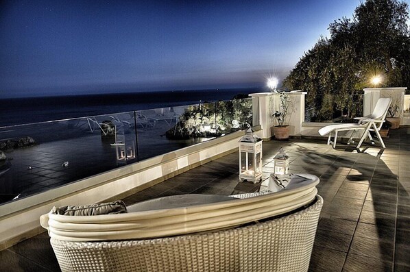 Una terraza en la azotea con piscina, sillas de descanso y vistas al océano de noche.
