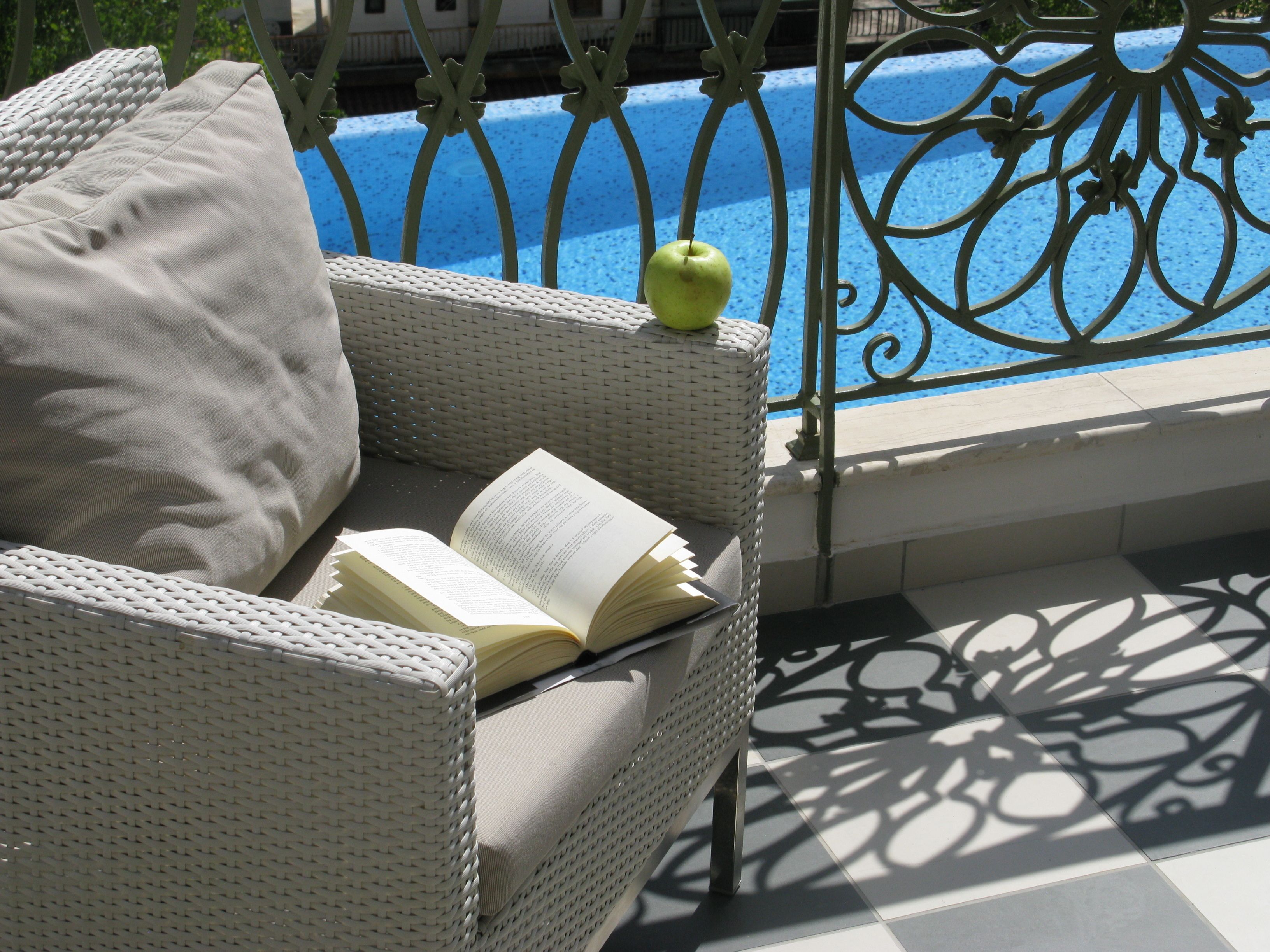 A wicker chair with a cushion and an open book, a green apple on the chair, and a pool with a metal fence in the background.