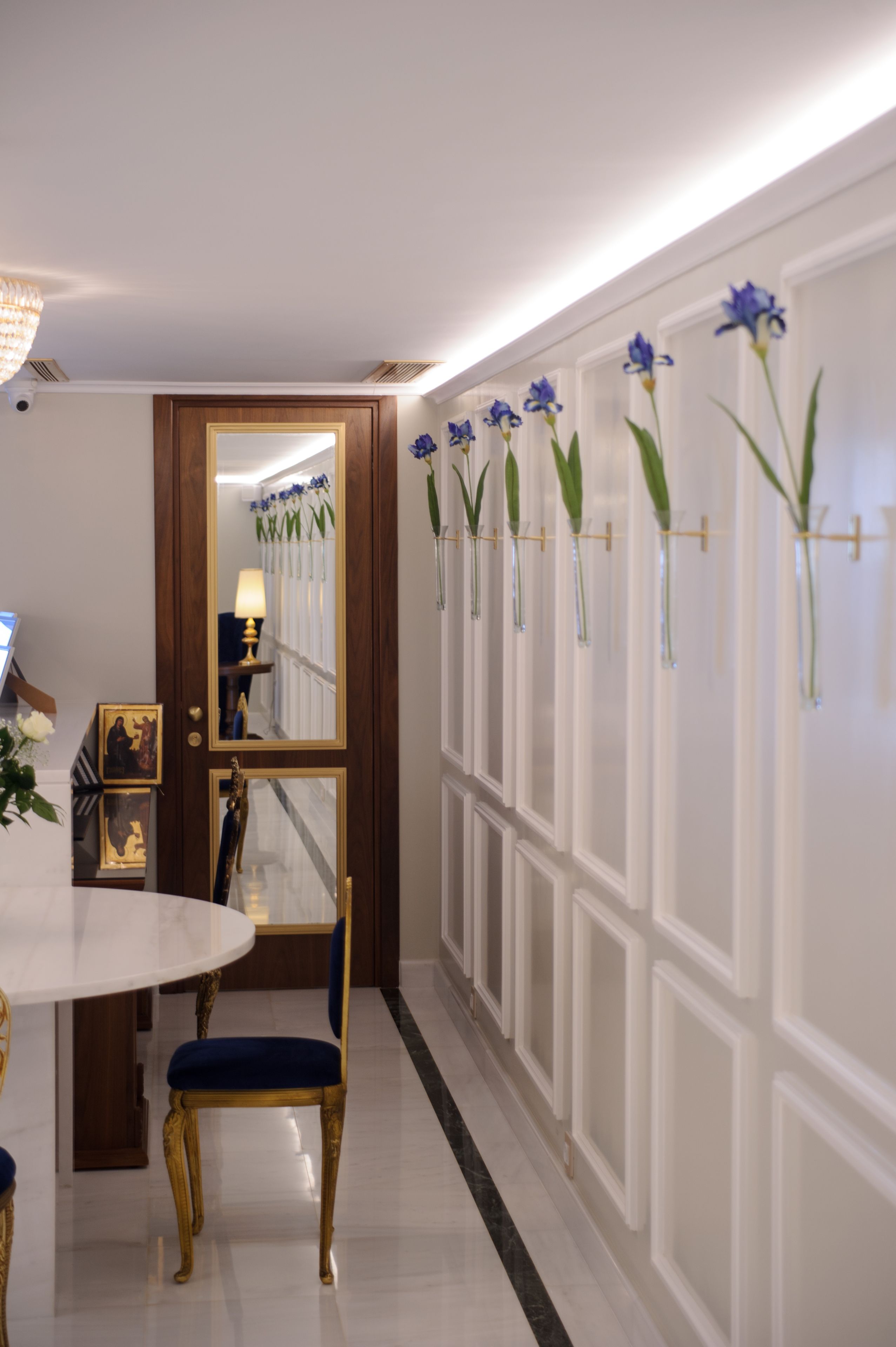 A hallway with a white wall, a wooden door, a round table with a blue chair, and a row of vases with blue flowers.