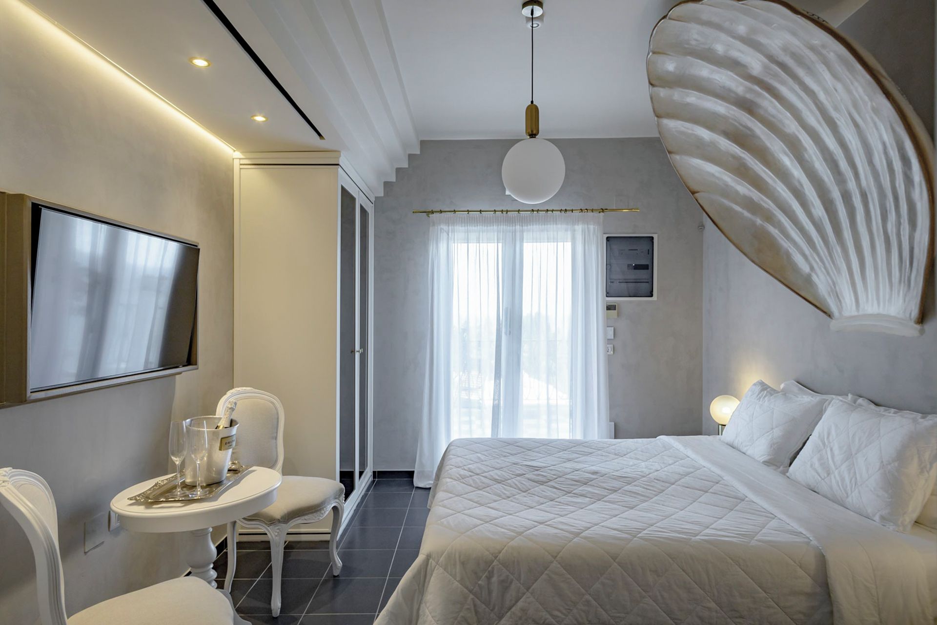 A modern hotel room with a large bed, a flat-screen TV mounted on the wall, a small table with a glass of water, and a decorative shell on the wall.