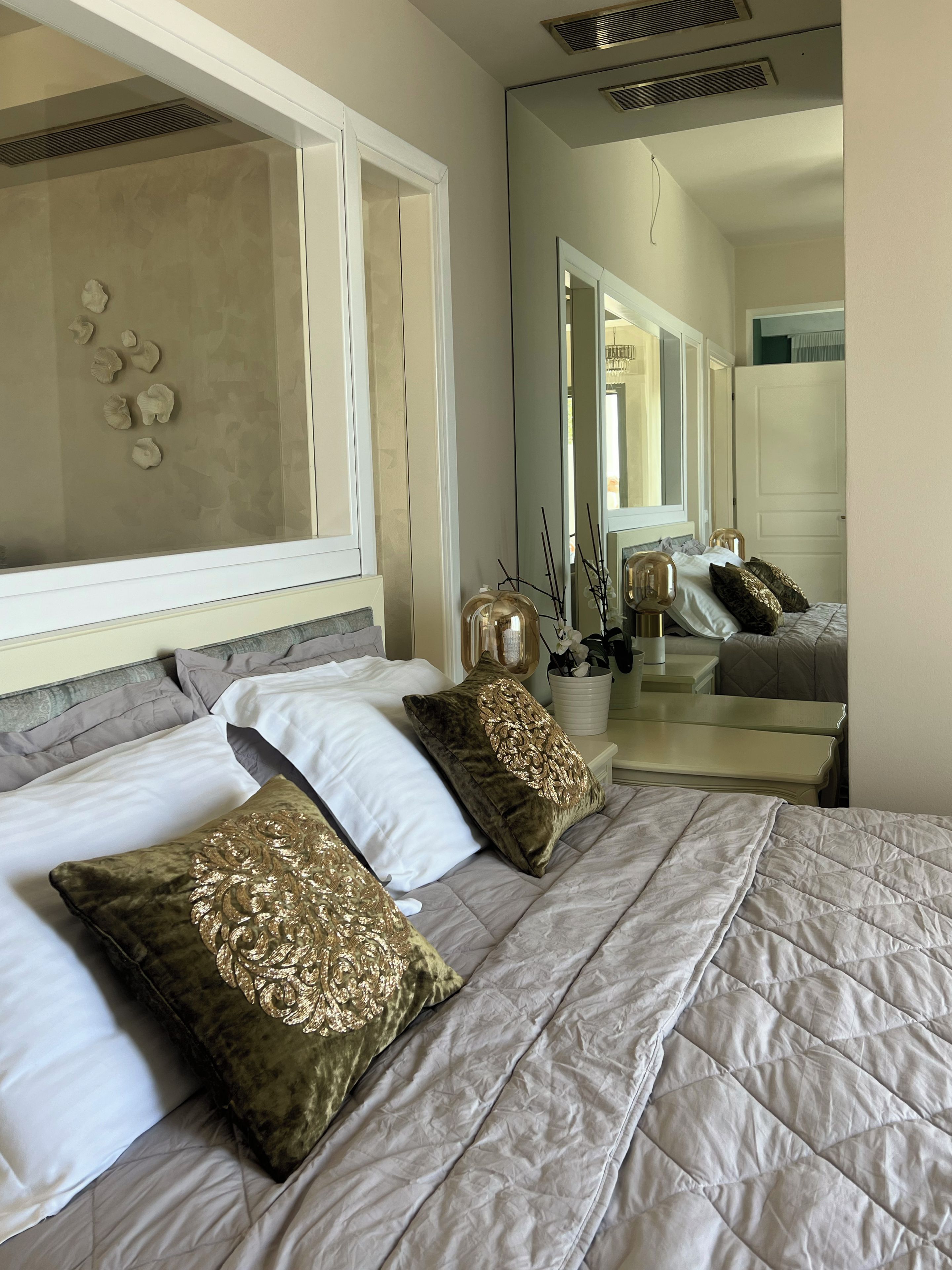 A neatly made bed with a quilted cover and decorative pillows, a bedside table with a vase and a lamp, and a large mirror reflecting the room.