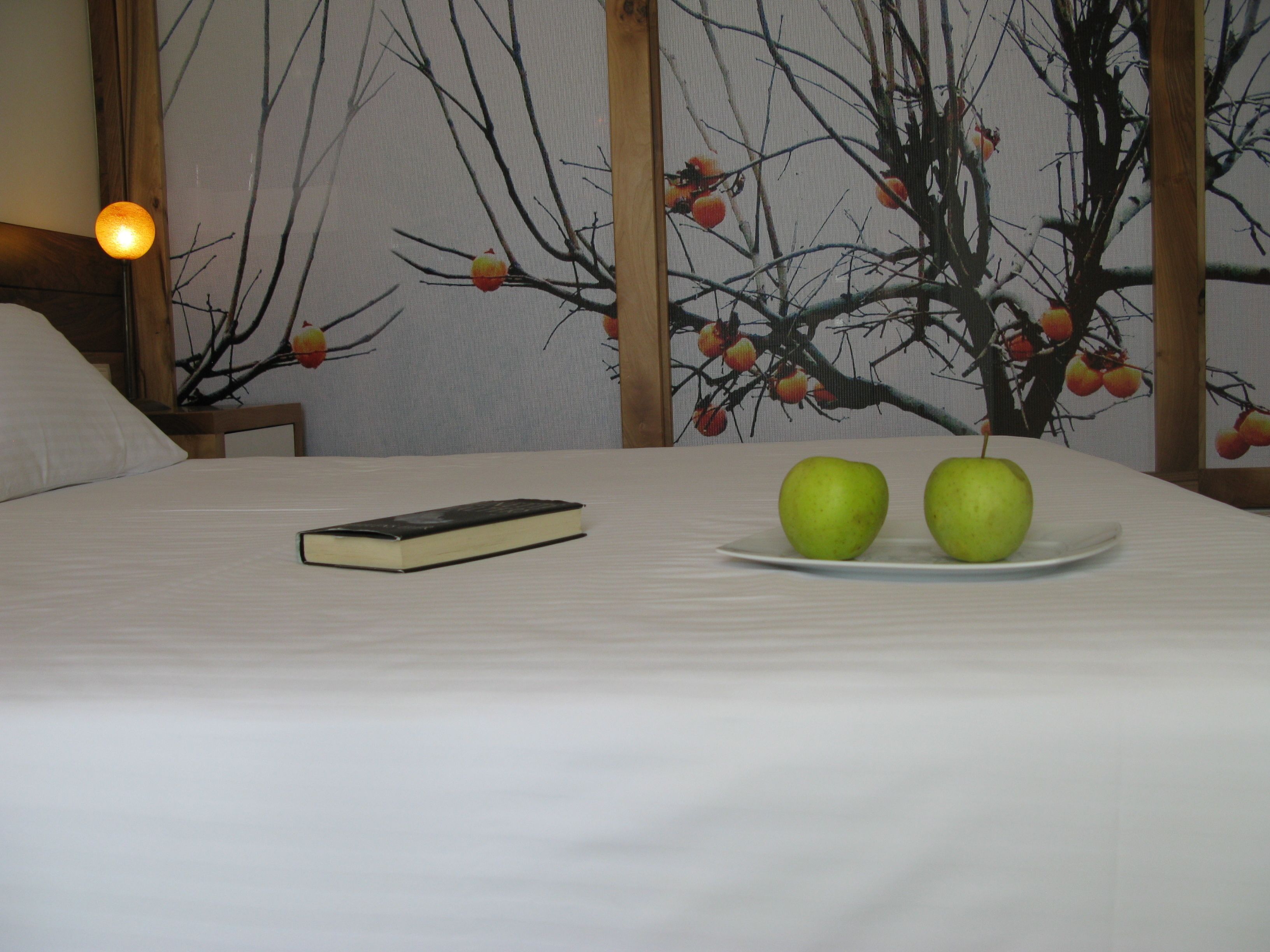 A neatly made bed with a book and two apples on a plate.