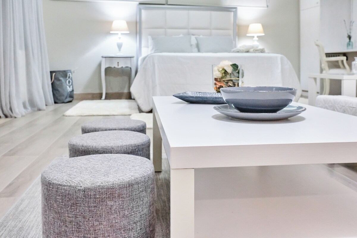 A modern hotel room with a white bed, a bedside table with a lamp, a small table with a bowl and a vase, and grey upholstered ottomans.