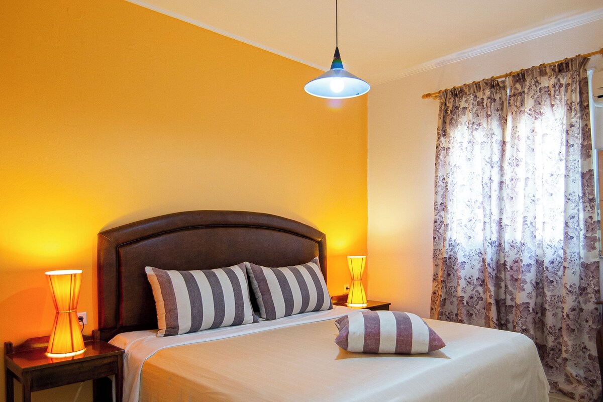 A neatly made bed with striped pillows, a bedside lamp, and a ceiling light fixture in a room with yellow walls and a window with floral curtains.