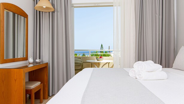 Habitación de hotel con cama, escritorio, silla, mesa con cesta y vistas al mar a través de amplias ventanas.