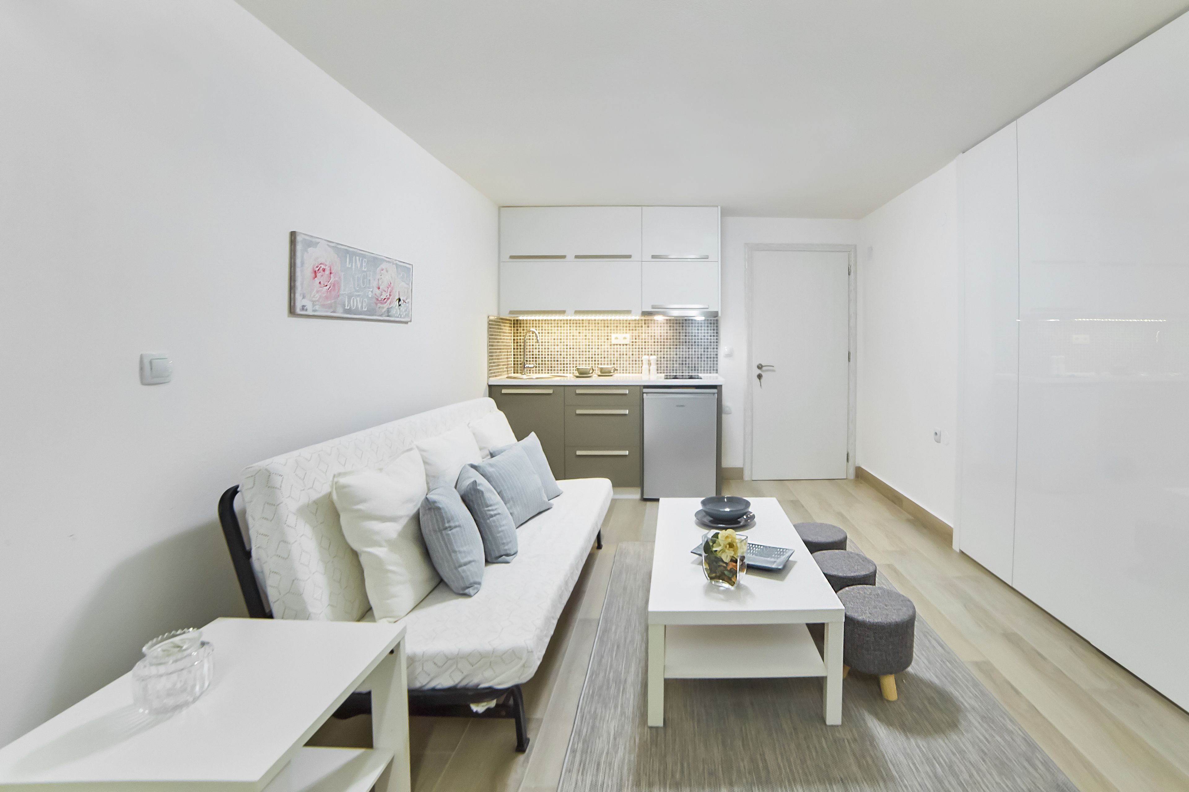 A modern apartment with a white sofa, a dining table, a kitchen area, and a small seating area with stools.