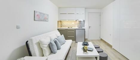 A modern apartment with a white sofa, a dining table, a kitchen area, and a small seating area with stools.