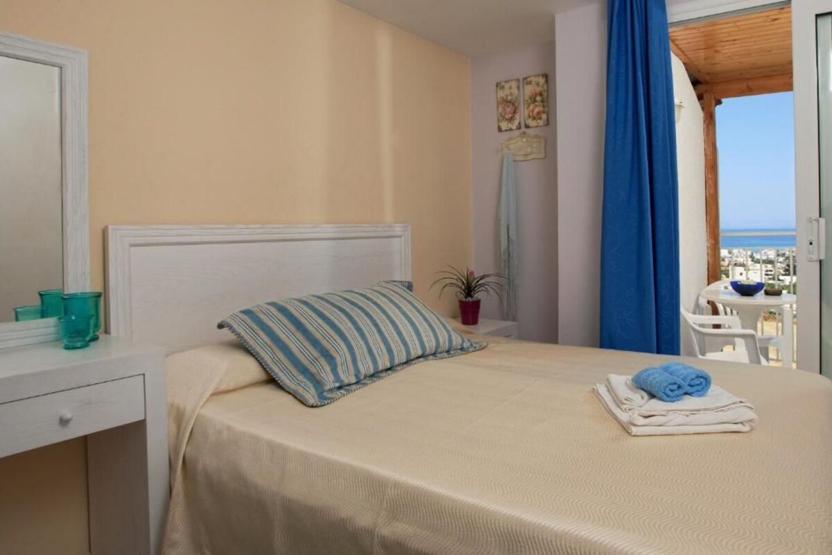 A hotel room with a bed, a nightstand, a potted plant, and a view of the sea through a balcony.