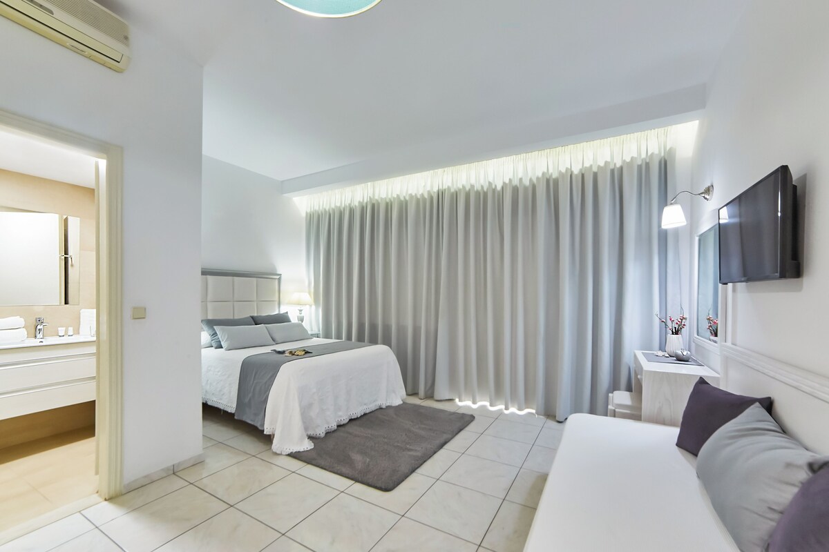 A modern hotel room with a large bed, a bedside table with a lamp, a TV mounted on the wall, and a bathroom with a mirror and sink.