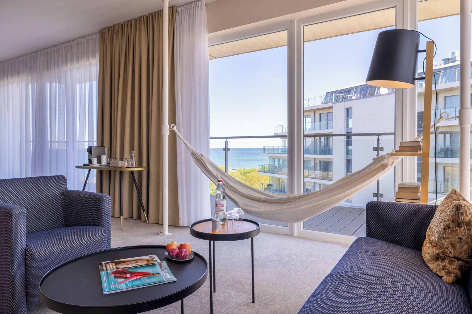 Habitación de hotel moderna con balcón grande, una hamaca, dos sofás, una mesa de centro y vista al mar.