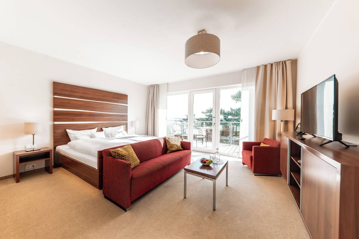 A hotel room with a bed, a red sofa, a small table, a TV, and a balcony with a view.
