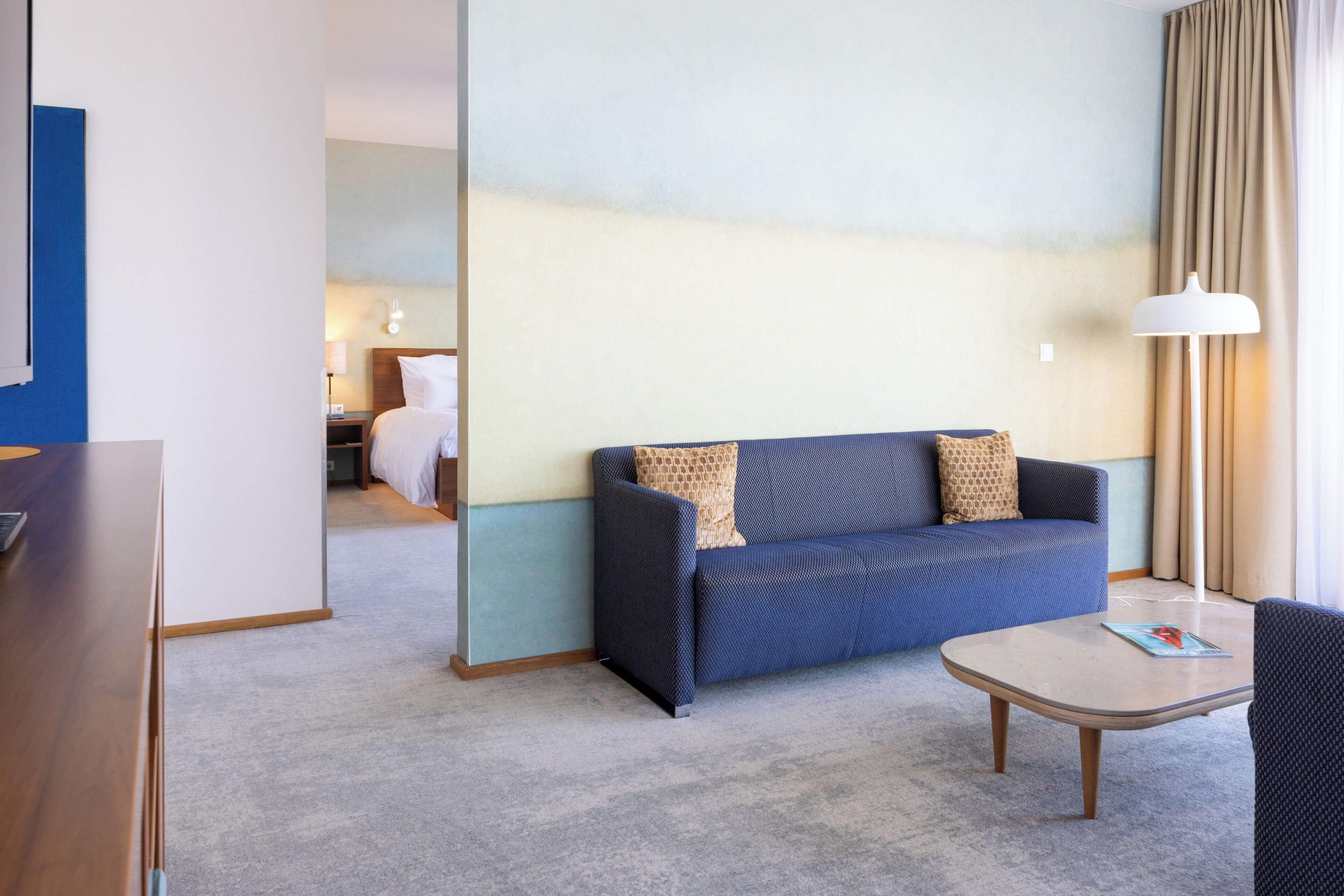 A hotel room with a sofa, a small table, a lamp, and a bed in the background.