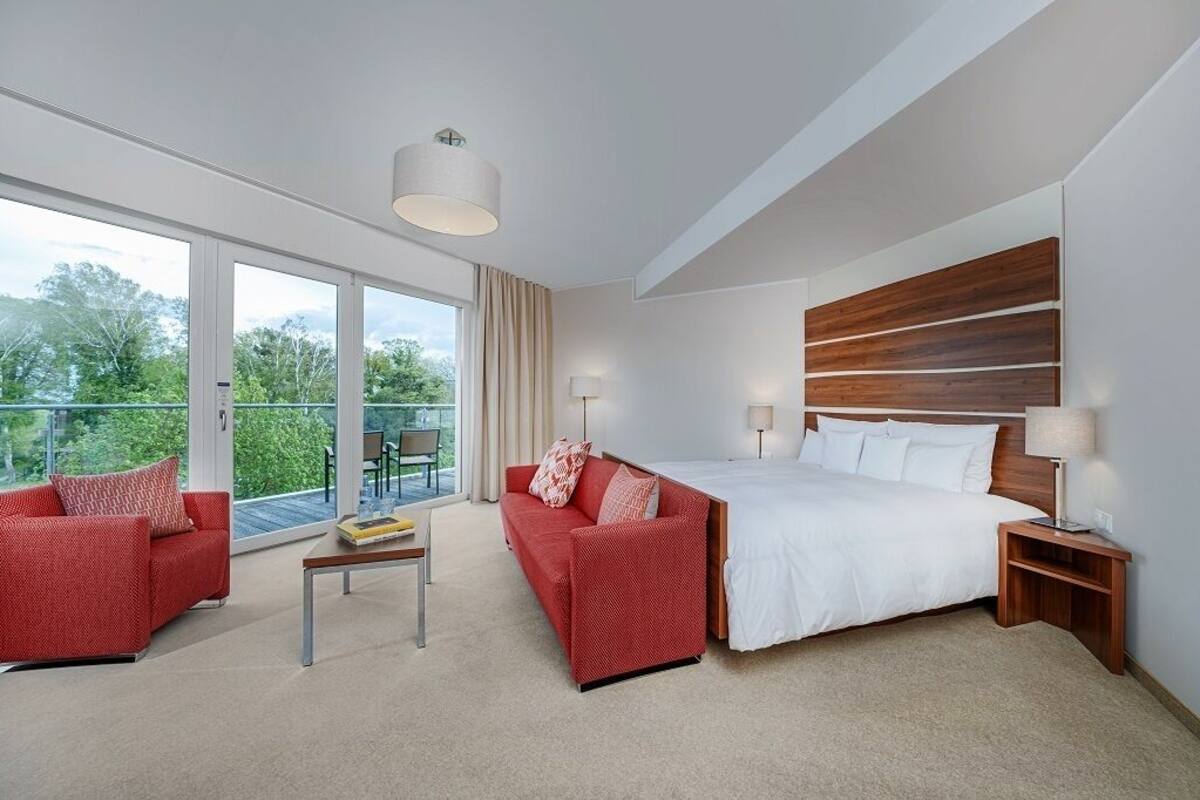 A modern hotel room with a large bed, two red sofas, a small table, and a balcony with a view of greenery.