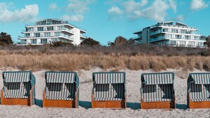 A beachfront property with a modern multi-story building, a sandy beach with striped beach chairs, and a grassy dune in the background. - THE GRAND Ahrenshoop (Ostseebad Ahrenshoop)