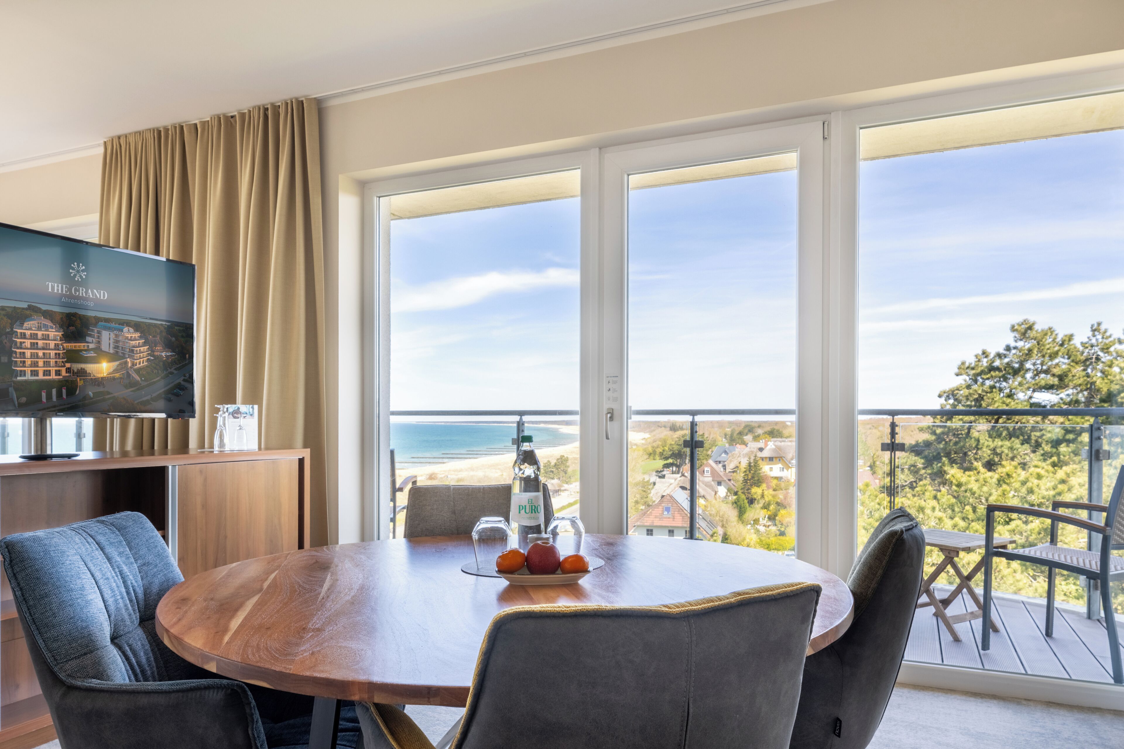 A hotel room with a balcony, a dining table with chairs, a TV, and a bottle of water.