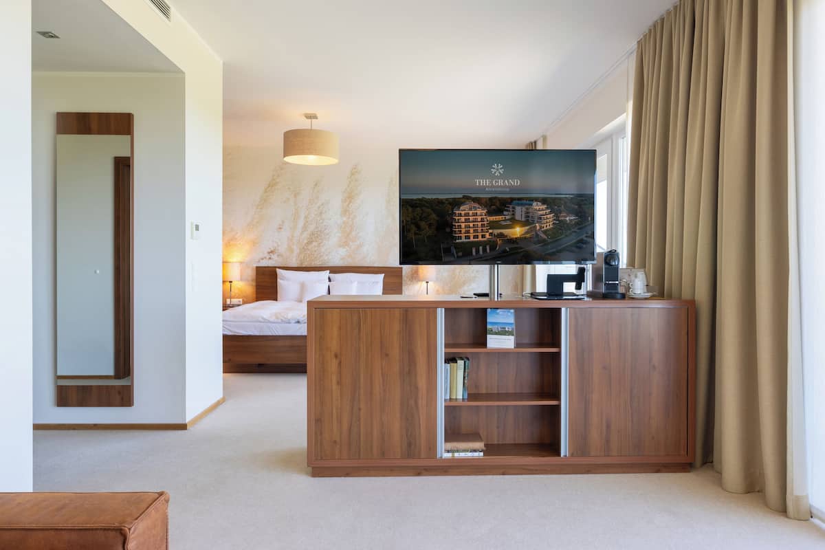 A hotel room with a bed, a TV mounted on a wooden cabinet, a bedside table with a lamp, and a view of the hotel exterior.