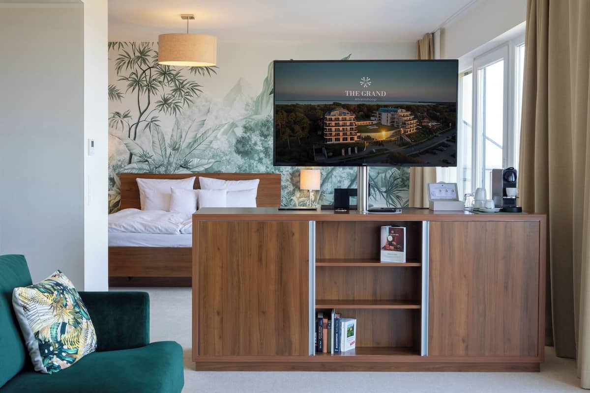 A hotel room with a bed, a TV mounted on a wooden cabinet, a green sofa, and a wall with a tropical mural.