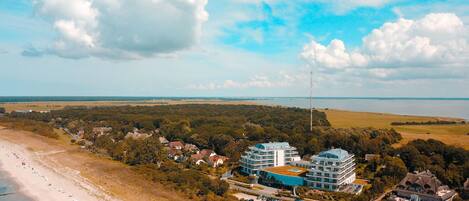 Luftaufnahme einer Küstenlandschaft mit Strand, einem großen Hotel mit mehreren Gebäuden und einer bewaldeten Landschaft im Hintergrund.