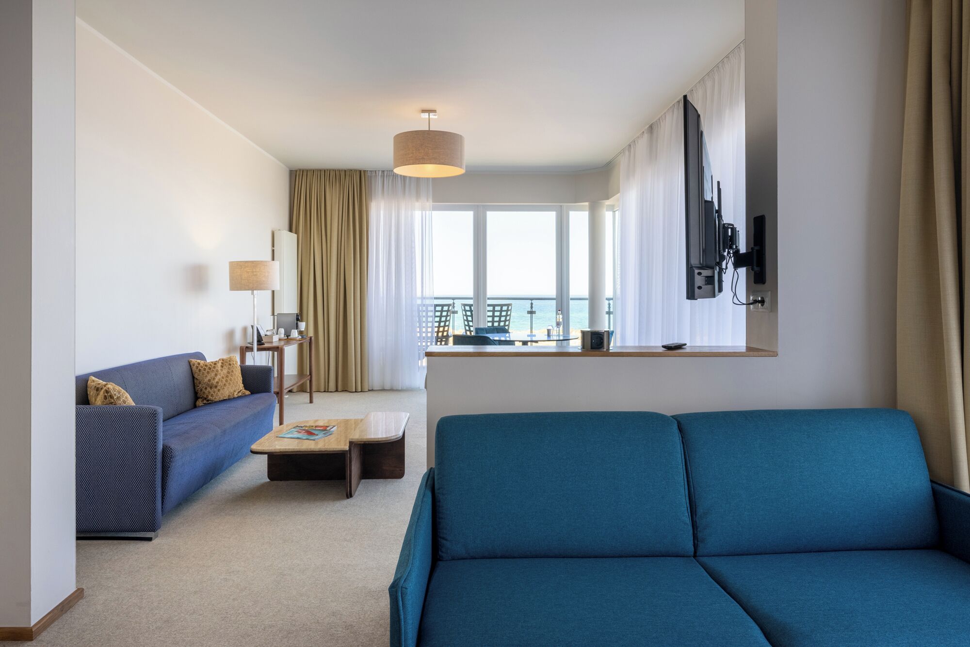 A modern hotel room with a blue sofa, a small table, a lamp, a TV mounted on the wall, and a view of the sea through large windows.