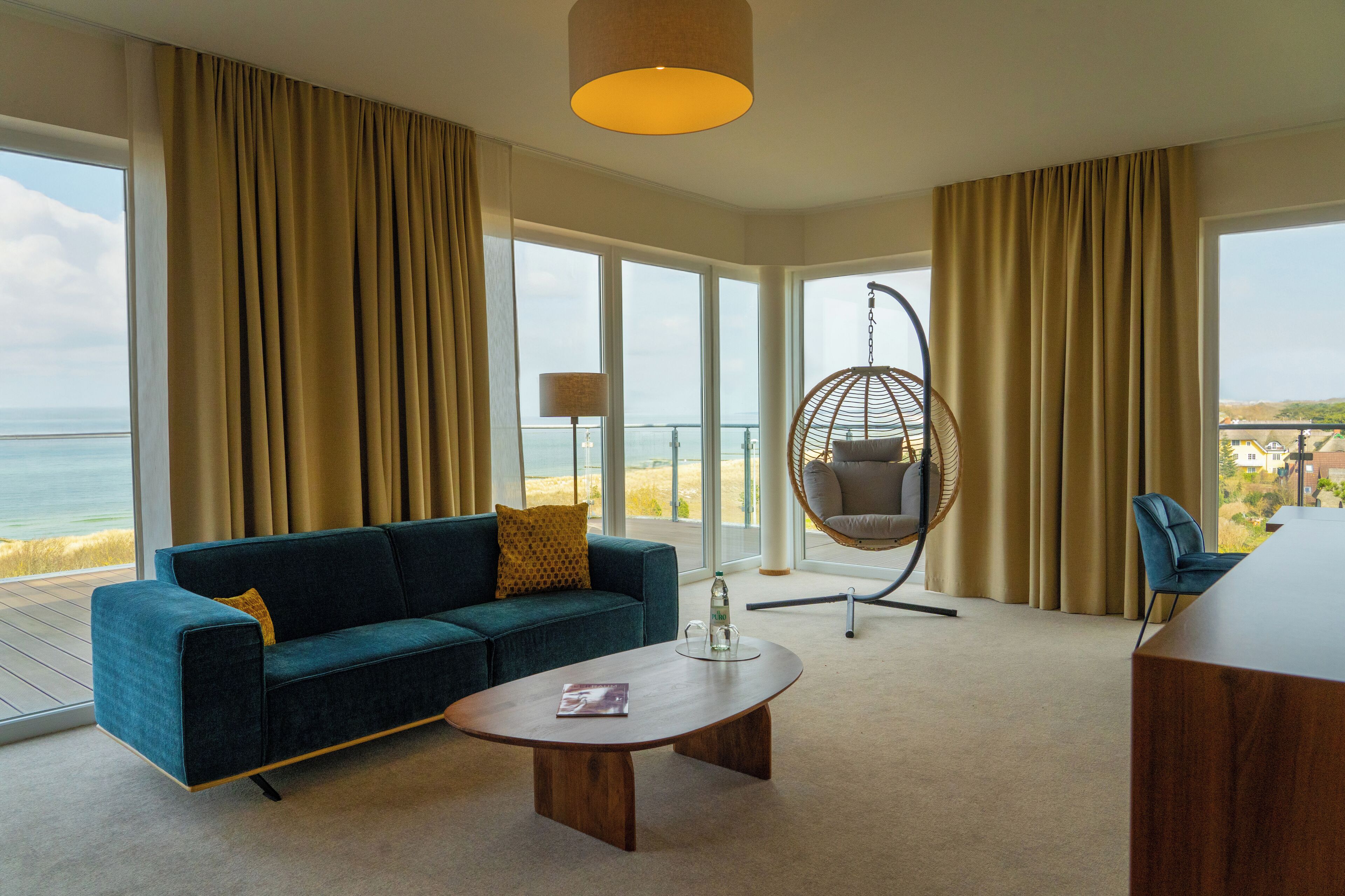 Une pièce moderne comprenant un canapé, une table basse, un fauteuil suspendu et de grandes fenêtres donnant sur la mer.
