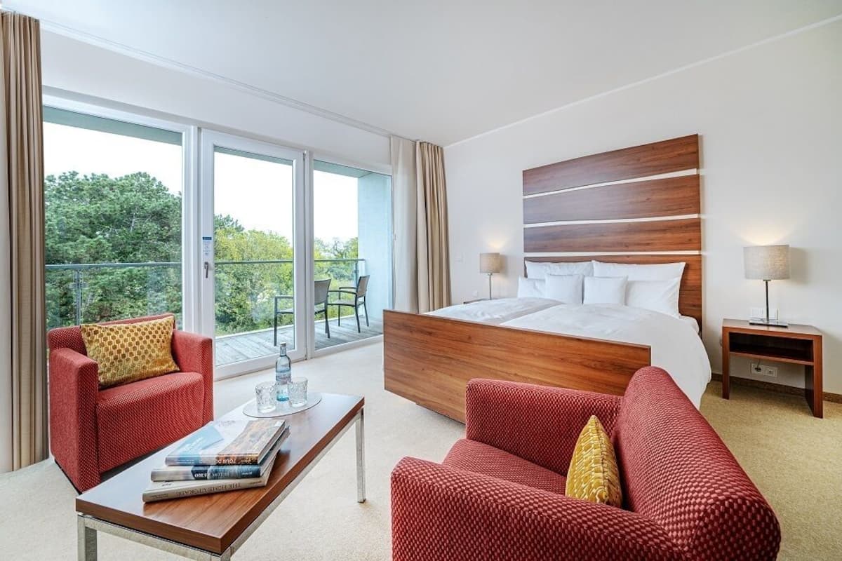 A modern hotel room with a large bed, two red sofas, a wooden coffee table, and a balcony with a view of trees.