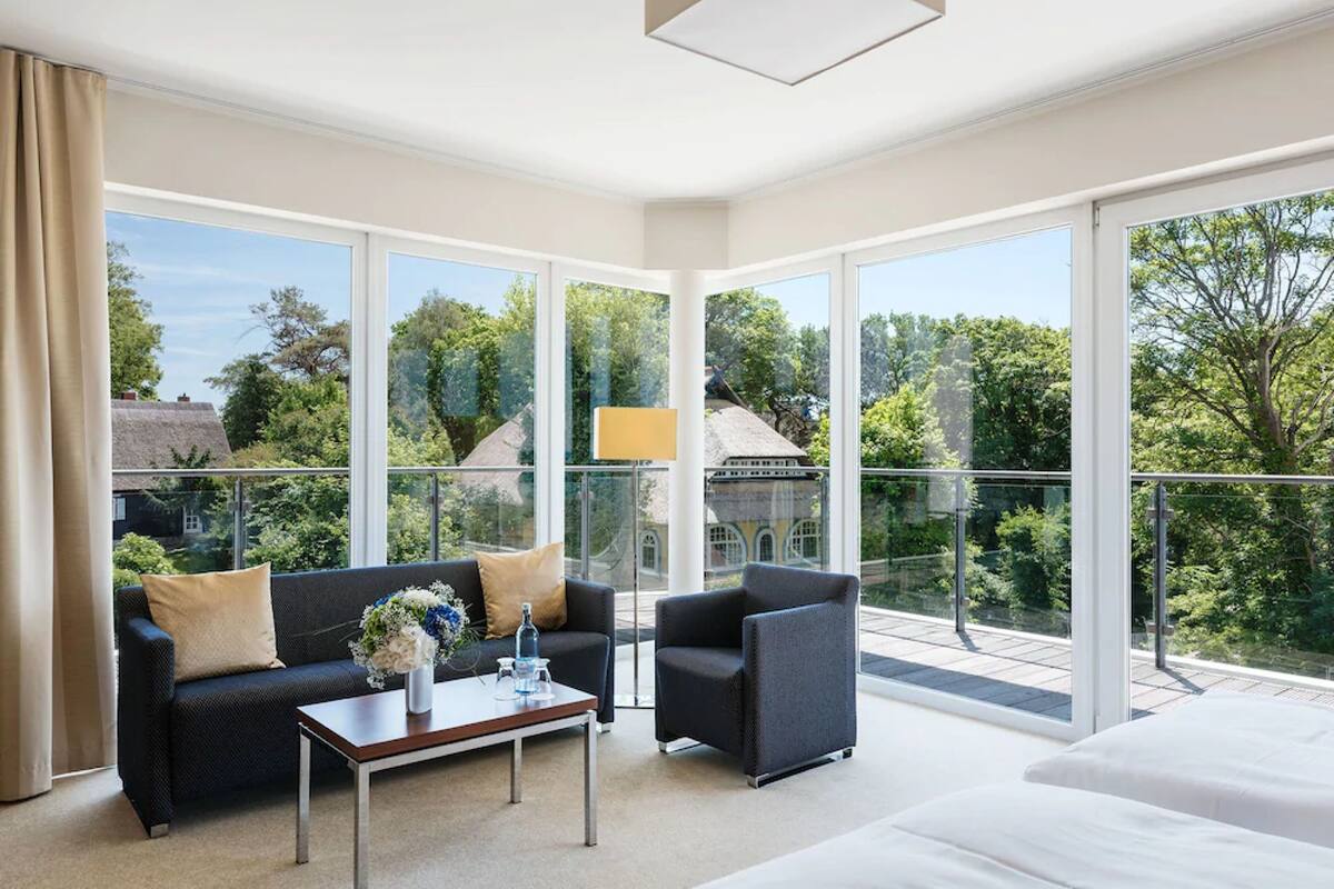 A modern hotel room with a large balcony, a sofa, a chair, a coffee table, and a bed.
