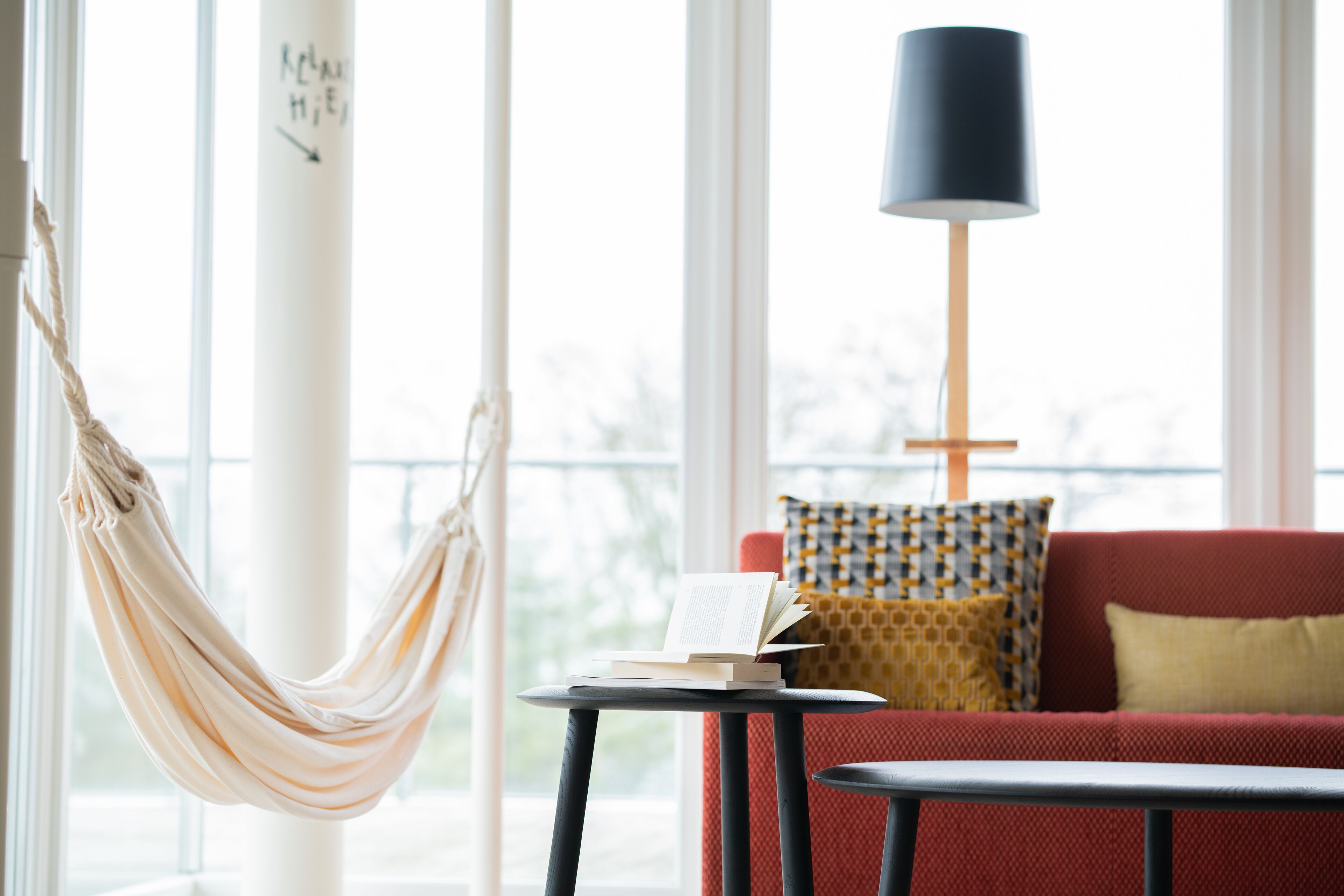 Et værelse med hængekøje, rød sofa, mønstret pude, lampe og et lille bord med en bog.