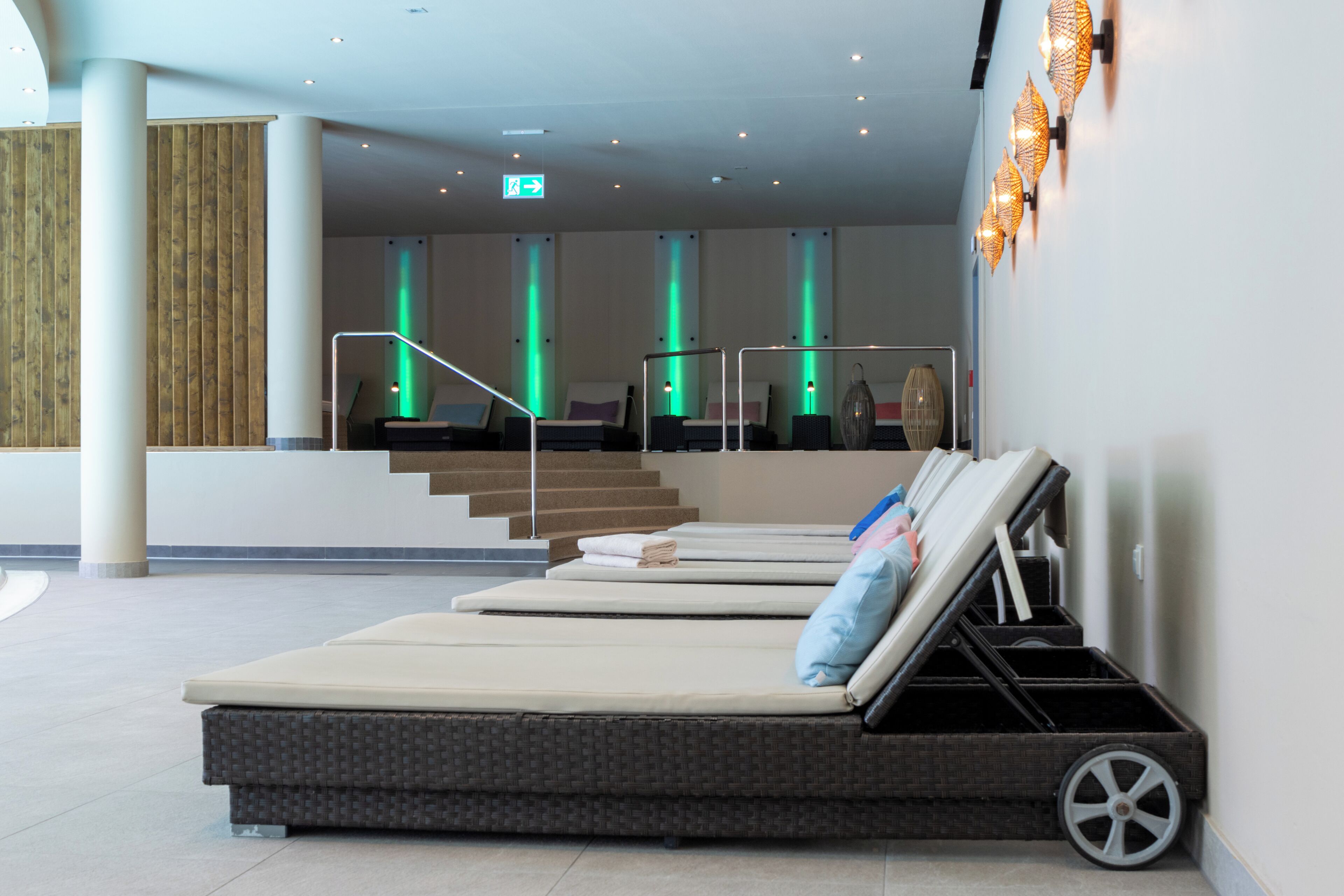 A modern hotel lobby with a reception desk, a staircase, and a lounge area with reclining chairs and a small cart.