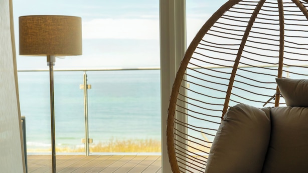 A wicker chair with cushions, a floor lamp, and a view of the sea through a window. - THE GRAND Ahrenshoop (Ostseebad Ahrenshoop)