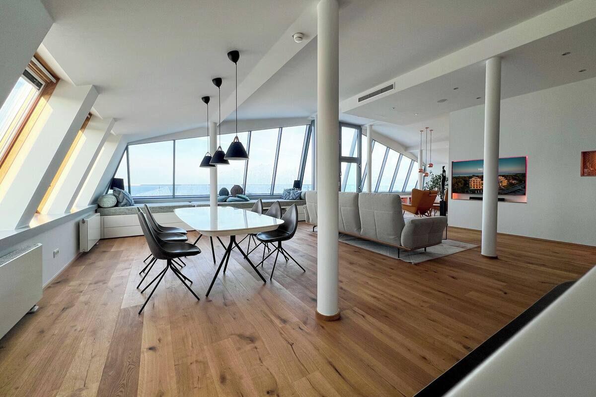 A spacious living area with a large round table, comfortable seating, and large windows offering a view of the sea.