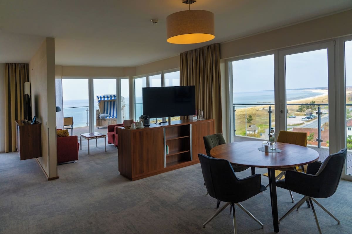 A spacious room with a large window, a flat-screen TV, a wooden cabinet, and a round dining table with chairs.