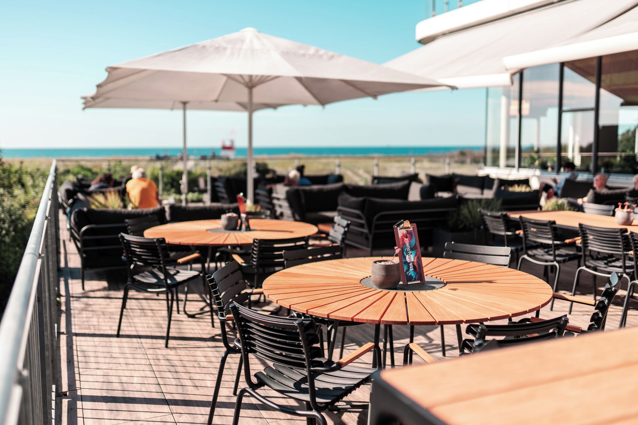 Eine Terrasse mit runden Holztischen und schwarzen Stühlen mit Blick auf einen Strand.