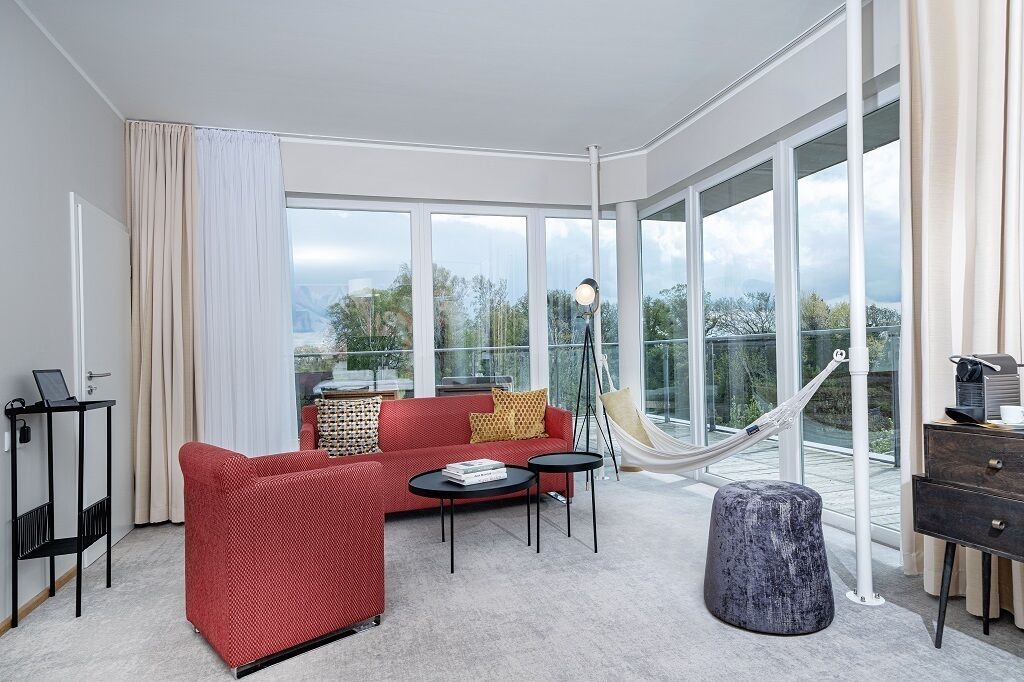 A modern hotel room with a red sofa, a small table, a hammock, and a view of the outdoors through large windows.