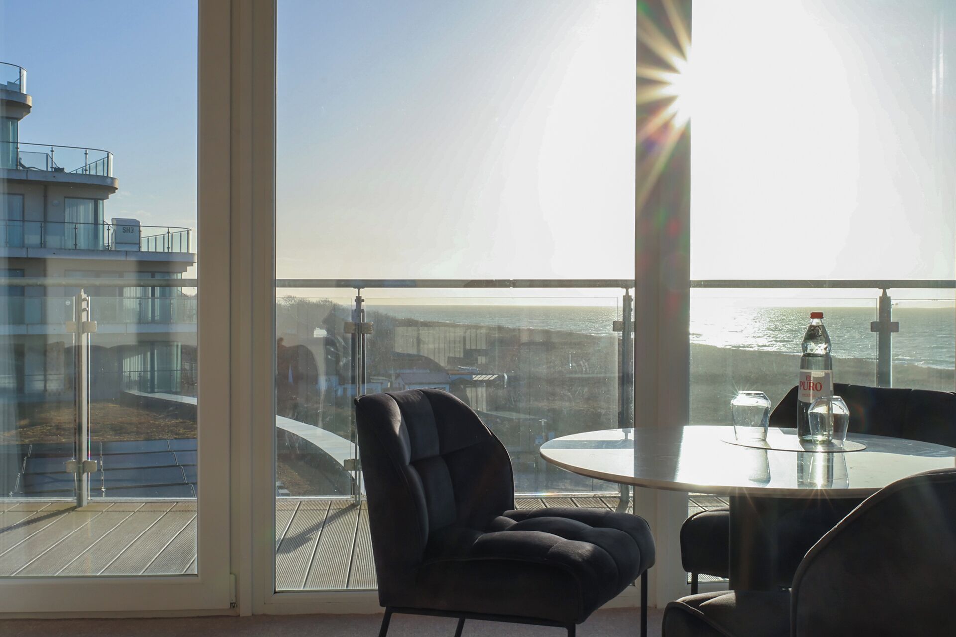 Un balcon avec une table et des chaises en verre, donnant sur une zone côtière avec des bâtiments et un ciel dégagé.