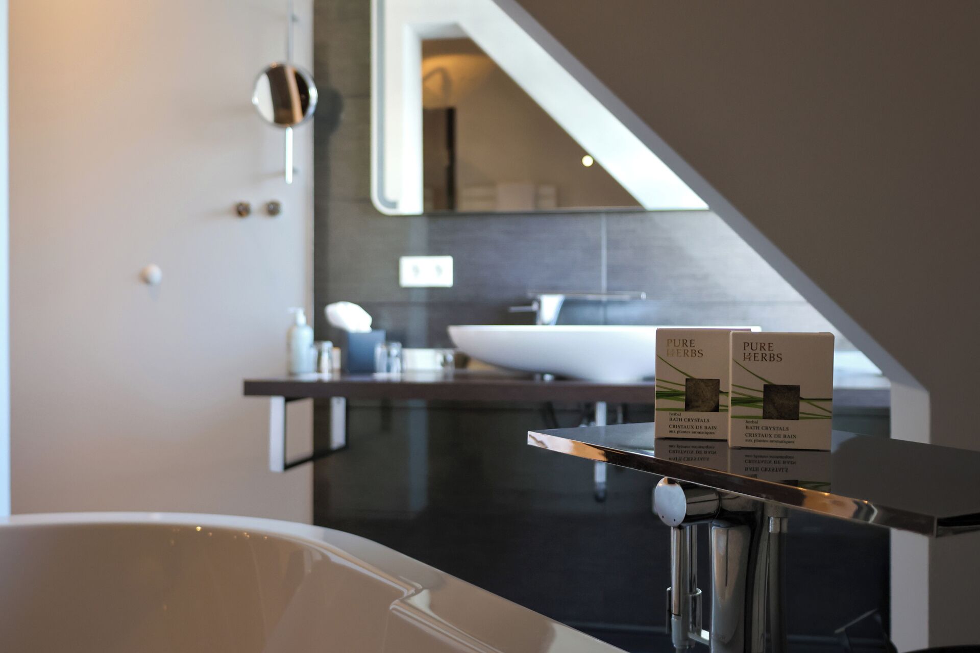 A modern bathroom with a bathtub, a sink, and a mirror. There are two boxes labeled "PURE HERBS" on a shelf.