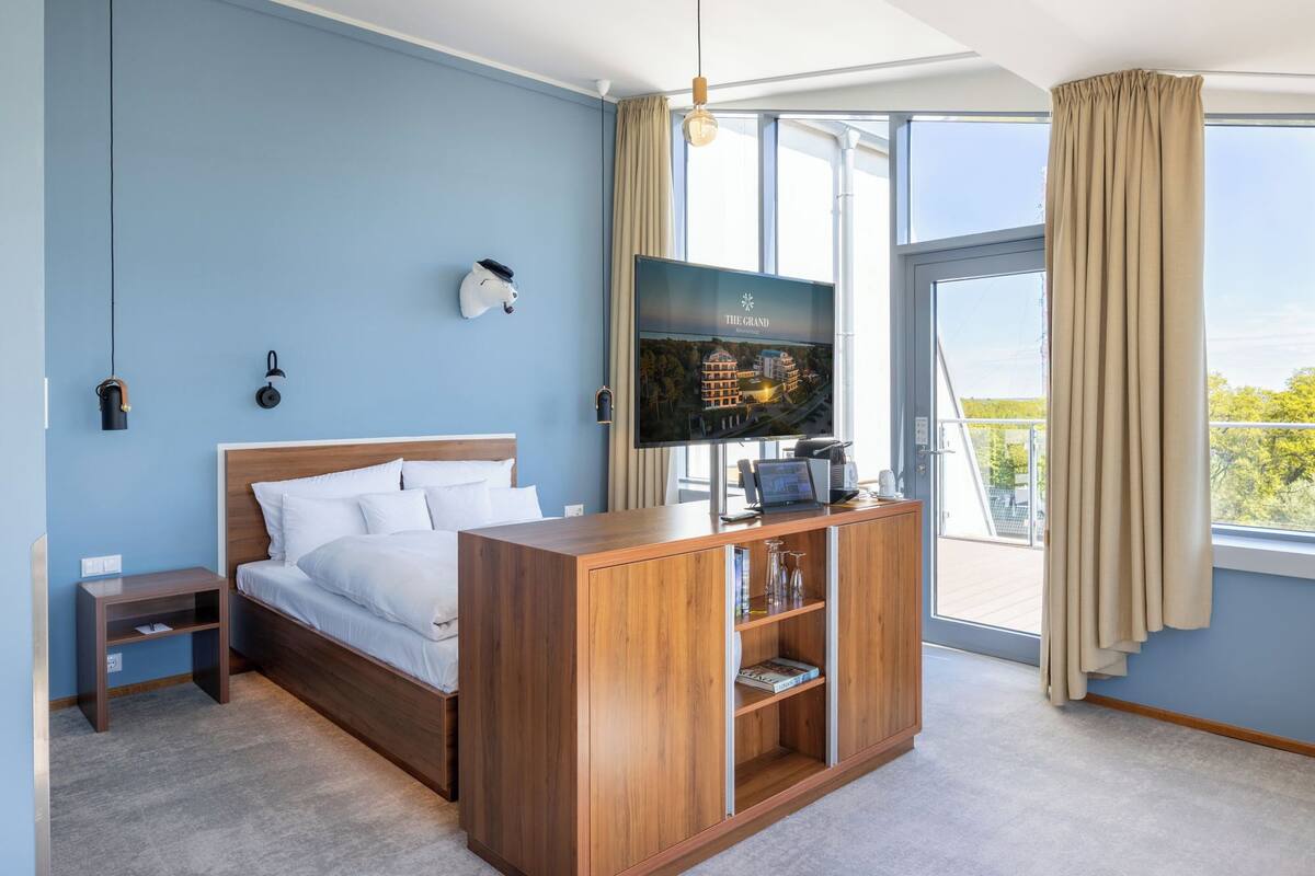 A hotel room with a bed, a TV, a wooden side table, and a large window with curtains.