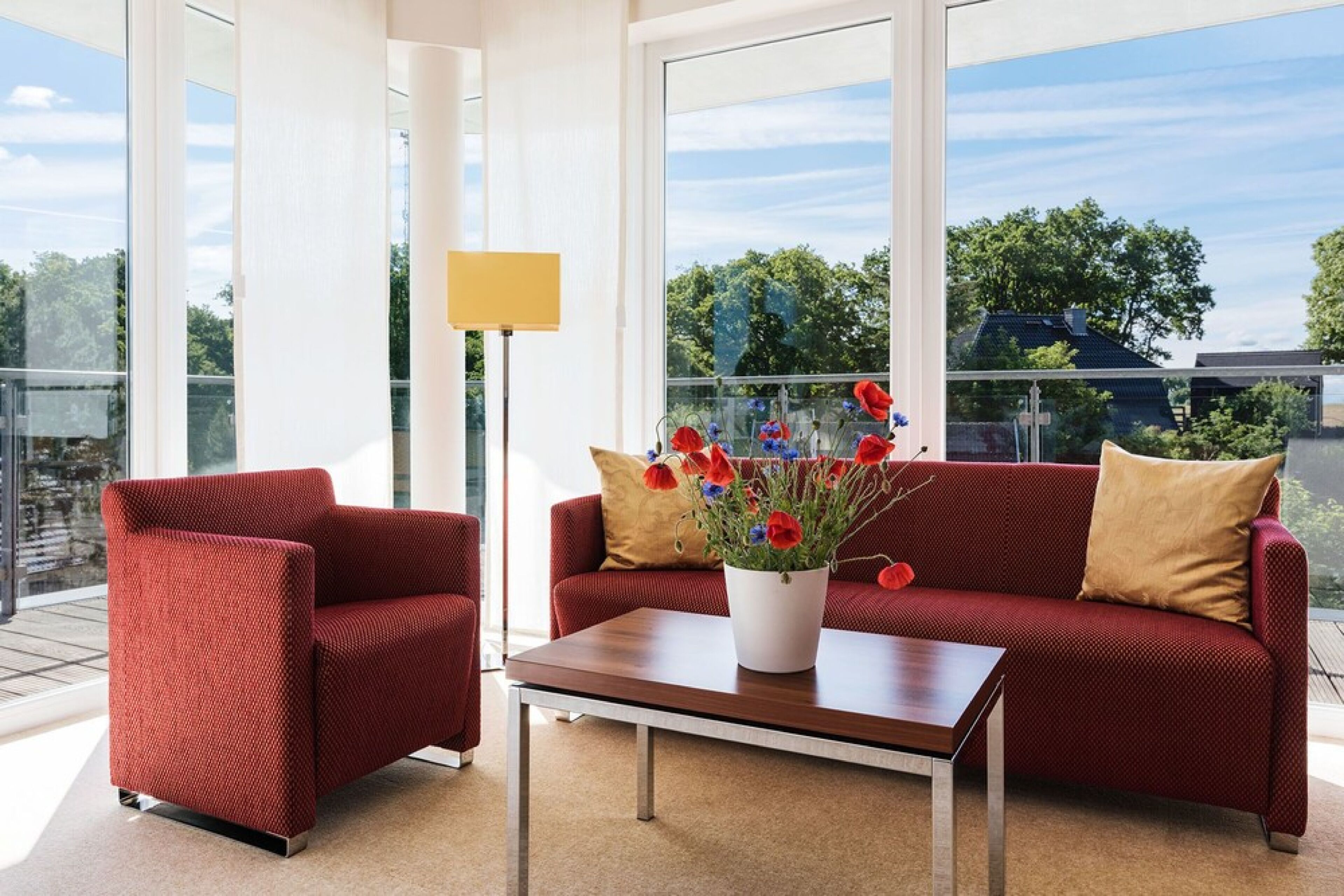 Una sala de estar moderna con un sofá rojo, un sillón a juego y una mesa de centro. Se ve una ventana grande con vistas a zonas verdes y edificios.