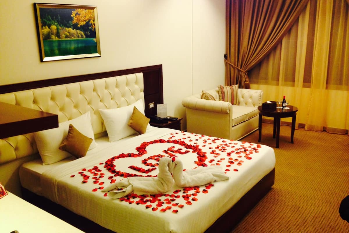 A hotel room with a bed adorned with rose petals and a white towel shaped like a heart. There is a painting on the wall, a sofa, and a small table with a bottle and glasses.