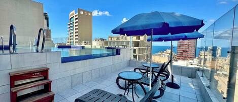 Un área de piscina en la azotea con vistas a la ciudad, piscina, sillas de descanso y un paraguas azul.