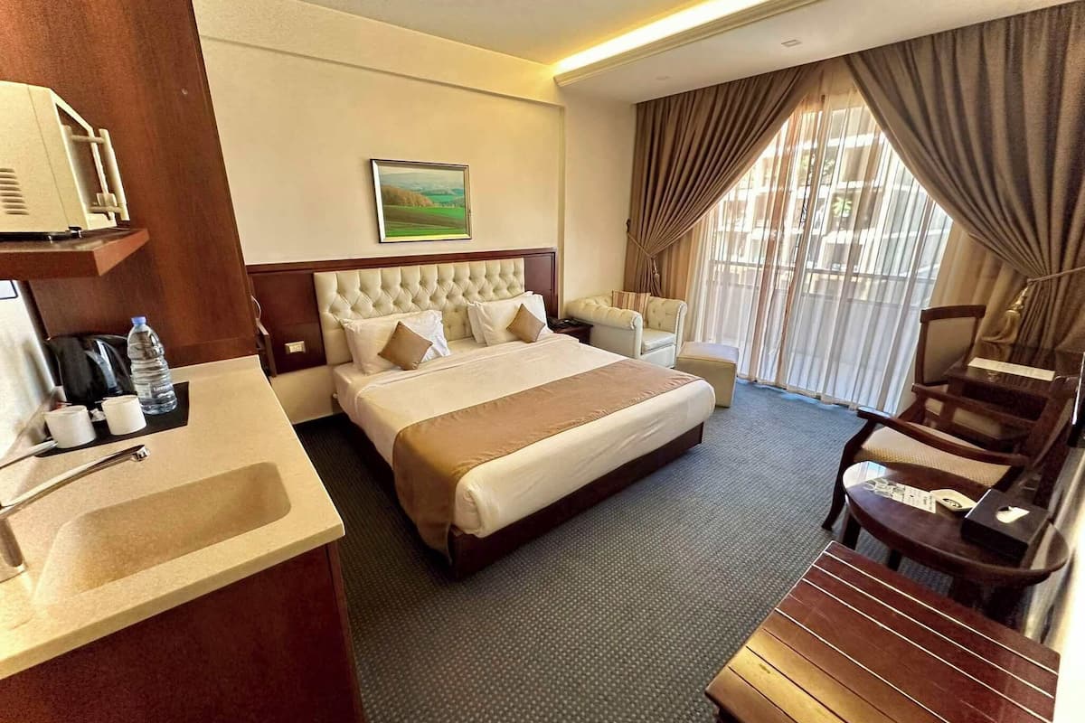A hotel room with a large bed, a bedside table with a lamp, a chair, a desk, and a window with curtains.