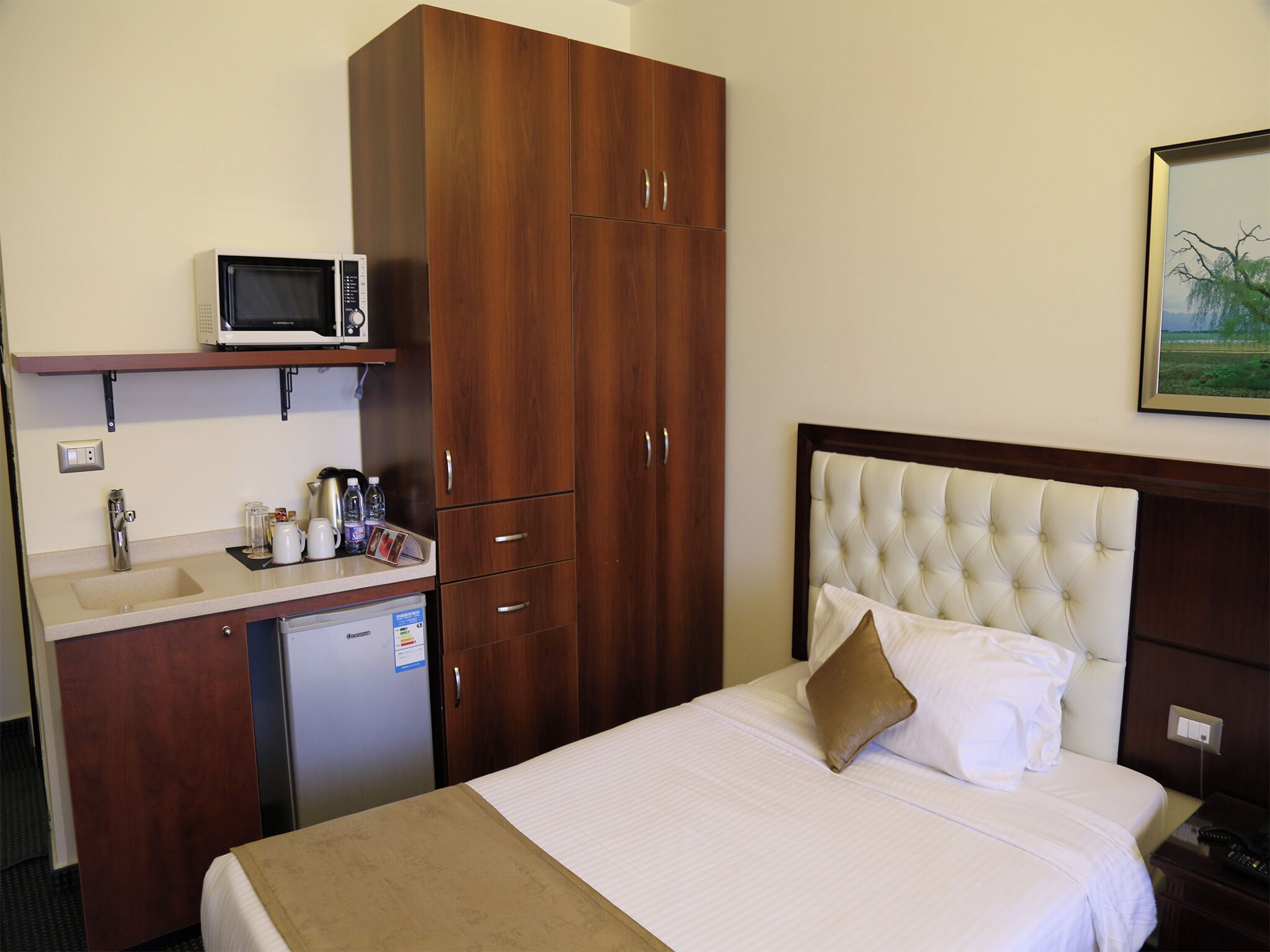 Una habitación de hotel con cama, un armario de madera, una kitchenette con microondas, un lavamanos y un refrigerador pequeño.