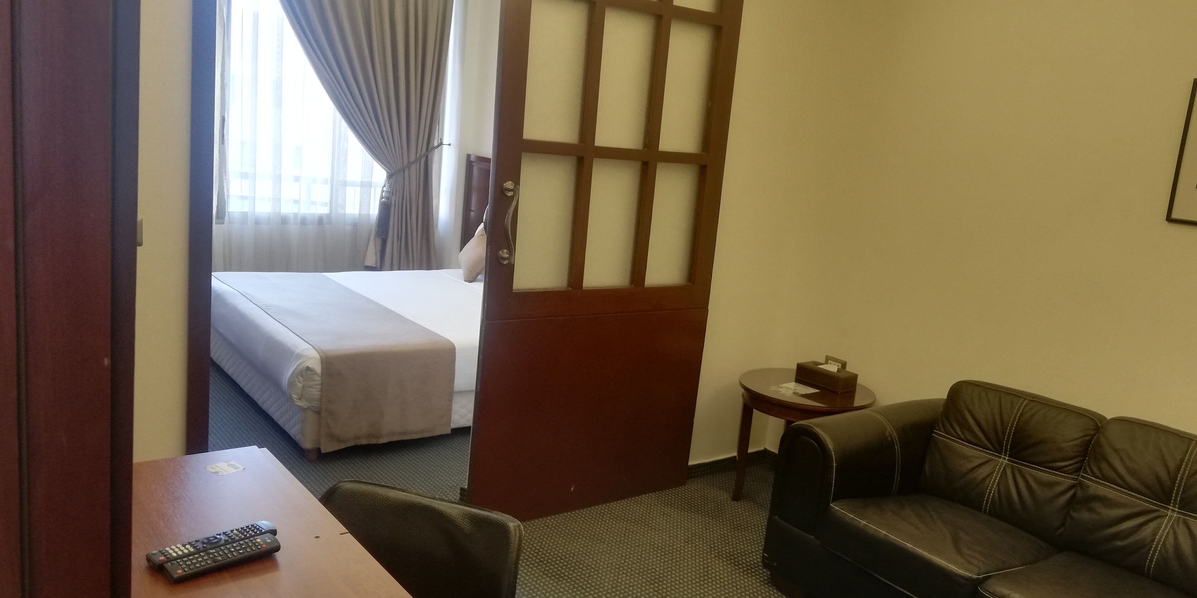 A hotel room with a bed, a leather sofa, a small table with a remote control, and a window with curtains.